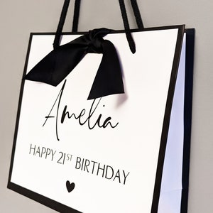 May include: White gift bag with black trim, handles, and bow. The bag features the name "Amelia" in cursive, the text "HAPPY 21ST BIRTHDAY," and a small heart symbol. A stylish and personalized gift bag.
