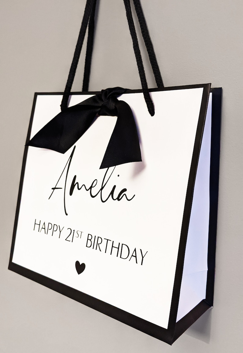 May include: White gift bag with black trim, handles, and bow. The bag features the name "Amelia" in cursive, the text "HAPPY 21ST BIRTHDAY," and a small heart symbol. A stylish and personalized gift bag.