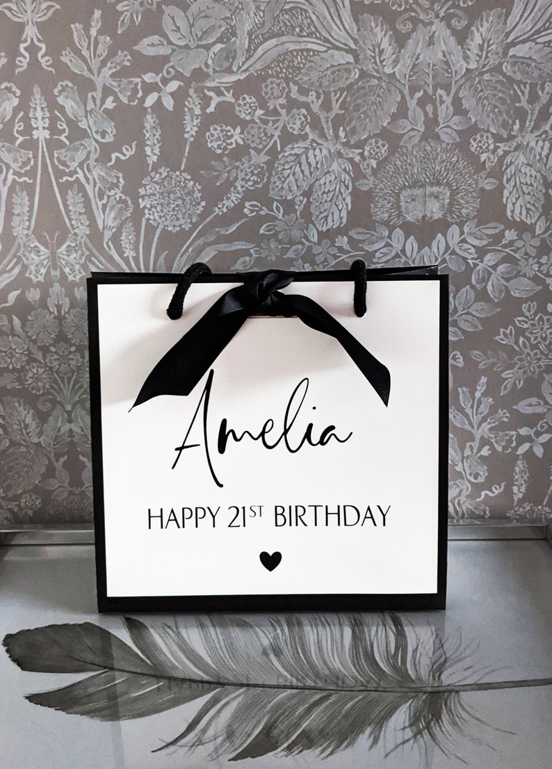 May include: A white gift bag with black trim and handles, tied with a black ribbon. The bag features the name "Amelia", the text "HAPPY 21ST BIRTHDAY", and a heart symbol in black.