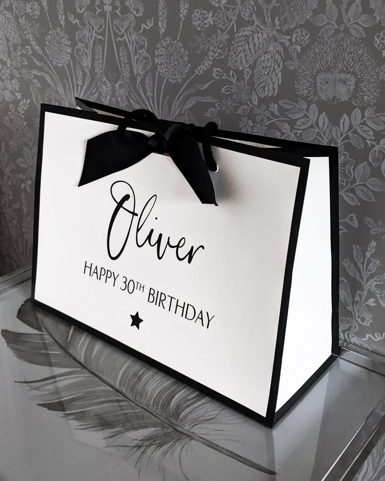 May include: A white gift bag with black trim and a black ribbon bow. The bag has the name "Oliver" in cursive, along with "Happy 30th Birthday" and a star graphic.