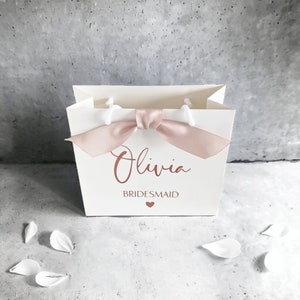 K&ouml;nnte beinhalten: Eine wei&szlig;e Geschenkt&uuml;te aus Papier mit einer Schleife aus altrosa Satinband an der Oberseite. Auf der Vorderseite ist der Name Olivia in Schreibschrift, das Wort BRIDESMAID in Blockbuchstaben und ein kleines Herz in Altrosa aufgedruckt.