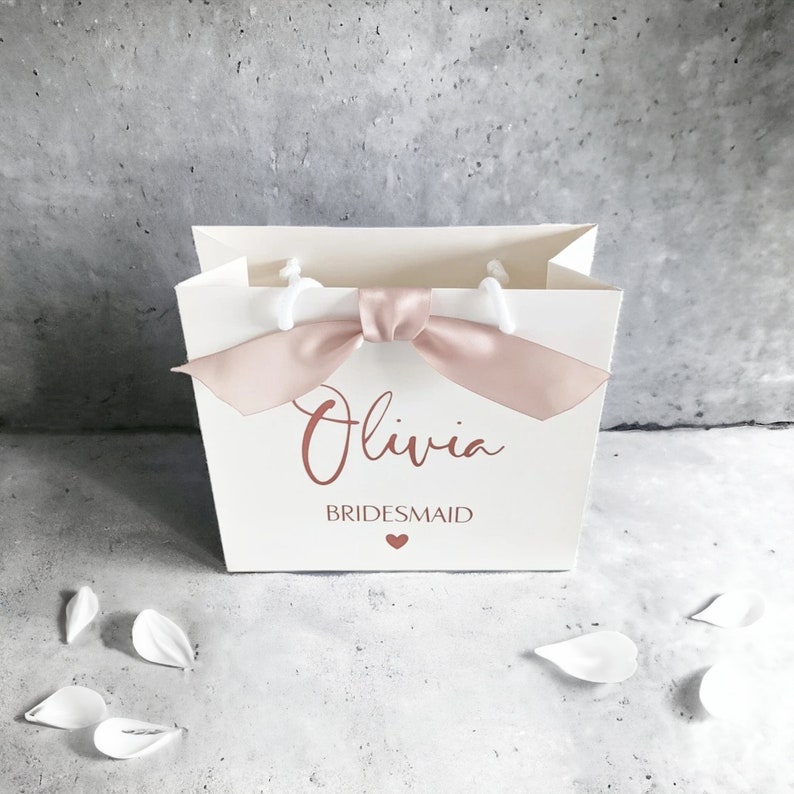 K&ouml;nnte beinhalten: Eine wei&szlig;e Geschenkt&uuml;te aus Papier mit einer Schleife aus altrosa Satinband an der Oberseite. Auf der Vorderseite ist der Name Olivia in Schreibschrift, das Wort BRIDESMAID in Blockbuchstaben und ein kleines Herz in Altrosa aufgedruckt.