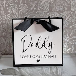 Może przedstawiać: Biała torba prezentowa z czarnymi uchwytami i czarną kokardką. Torba ma czarny napis "Daddy Love From Hannah" z czarnym sercem.