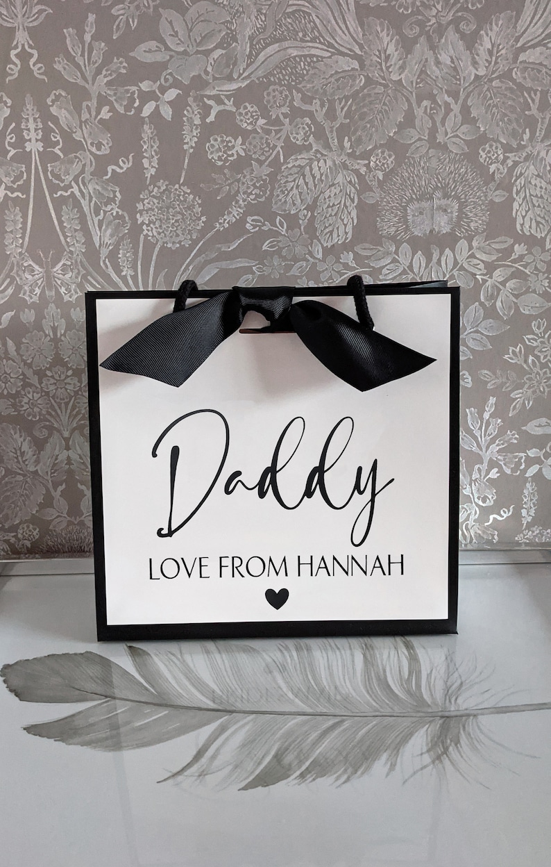 Może przedstawiać: Biała torba prezentowa z czarnymi uchwytami i czarną kokardką. Torba ma czarny napis "Daddy Love From Hannah" z czarnym sercem.