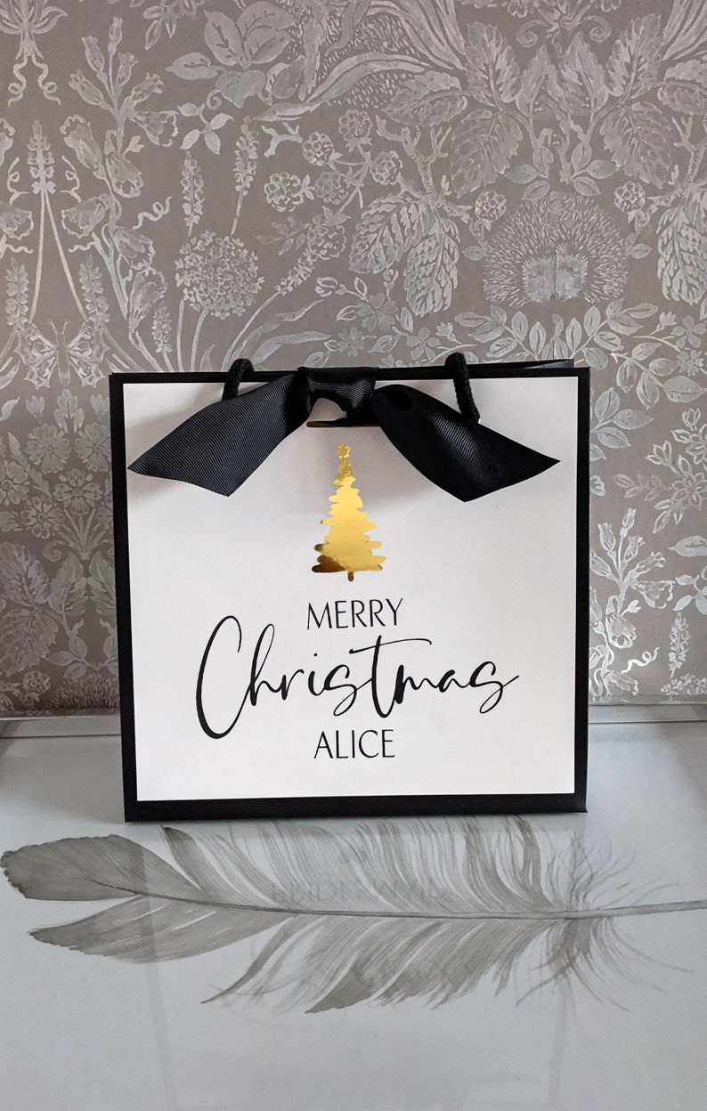 May include: A white gift bag with a black border and handles, featuring a gold Christmas tree graphic. The words "MERRY Christmas ALICE" are printed in black script. A black bow adorns the top.