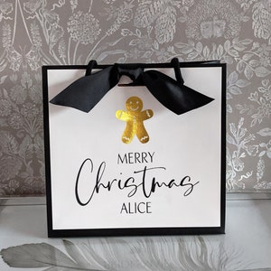 May include: A white gift bag with a black border and handles, featuring a gold gingerbread man and the text "MERRY Christmas ALICE" in black script. A black bow adorns the top. The bag is designed for Christmas gifting.