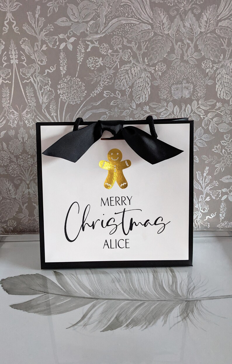 May include: A white gift bag with a black border and handles, featuring a gold gingerbread man and the text "MERRY Christmas ALICE" in black script. A black bow adorns the top. The bag is designed for Christmas gifting.