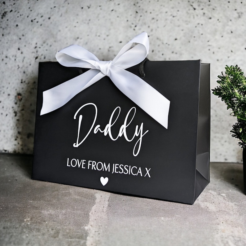 May include: Black gift bag with a white satin ribbon and bow. The bag has the word "Daddy" written in white cursive lettering and the text "Love from Jessica X" below it. A small white heart is printed below the text.