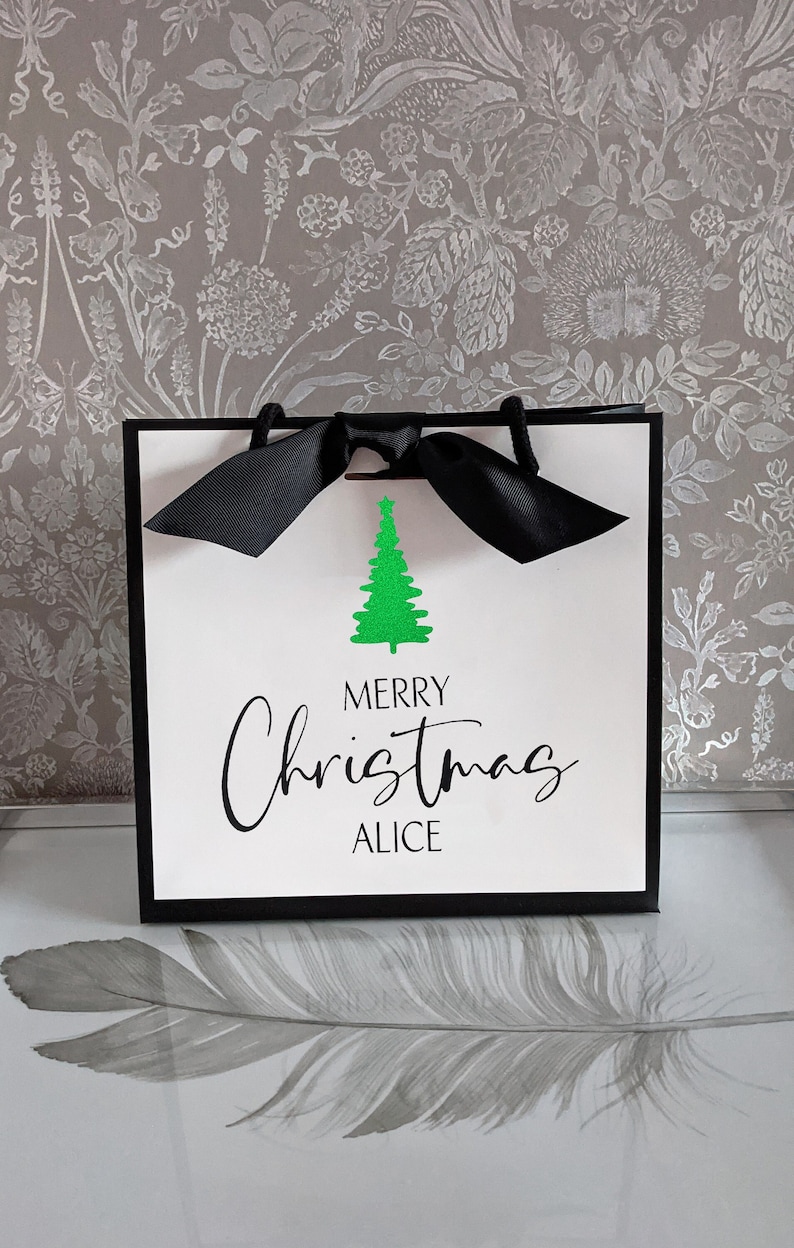 May include: A white gift bag with a black border and handles, tied with a black bow. The bag features a green glitter Christmas tree and the words "MERRY Christmas ALICE" in black script.