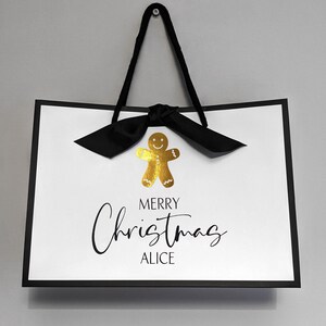 May include: White gift bag with black trim and handles, featuring a gold gingerbread man and a black bow. The text "MERRY Christmas ALICE" is written in black script, perfect for holiday gifting.
