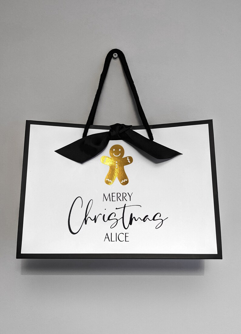 May include: White gift bag with black trim and handles, featuring a gold gingerbread man and a black bow. The text "MERRY Christmas ALICE" is written in black script, perfect for holiday gifting.