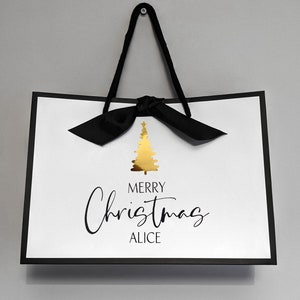 May include: White gift bag with a black border and handles, featuring a gold Christmas tree graphic. The text "MERRY Christmas ALICE" is written in black script. A black bow is tied at the top.