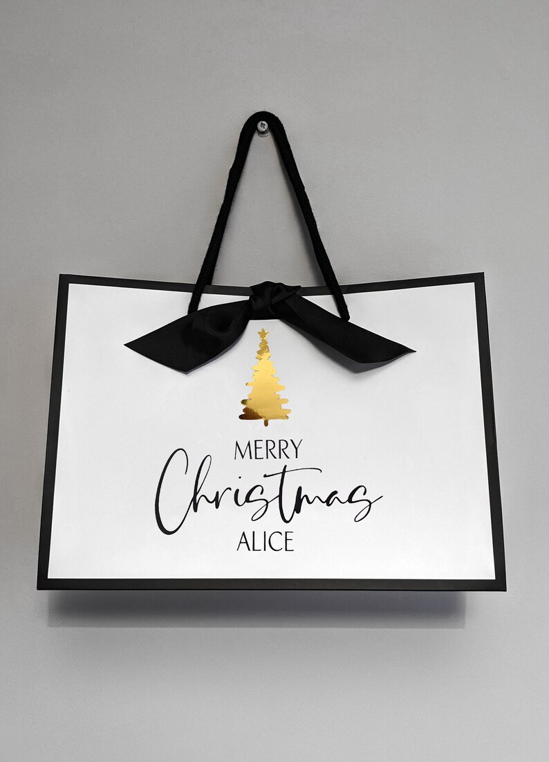 May include: White gift bag with a black border and handles, featuring a gold Christmas tree graphic. The text "MERRY Christmas ALICE" is written in black script. A black bow is tied at the top.