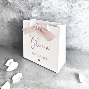 K&ouml;nnte beinhalten: Eine wei&szlig;e Geschenkt&uuml;te mit einem Griff aus altrosa Satinband. Auf der Vorderseite ist der Name Olivia in Schreibschrift aufgedruckt, gefolgt von dem Wort BRIDESMAID in serifenloser Schrift und einem kleinen Herzsymbol in passendem Altrosa.