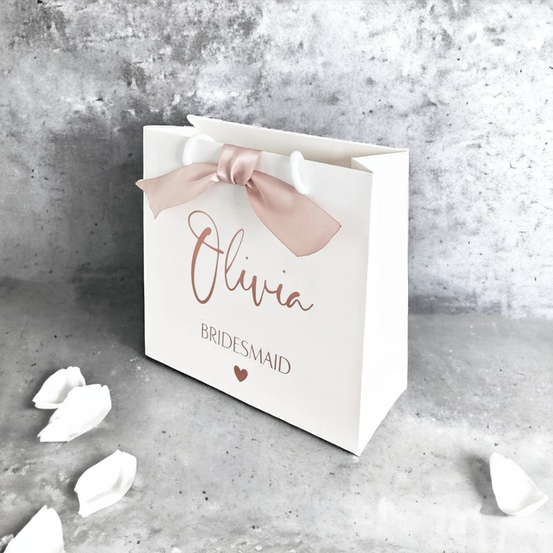 K&ouml;nnte beinhalten: Eine wei&szlig;e Geschenkt&uuml;te mit einem Griff aus altrosa Satinband. Auf der Vorderseite ist der Name Olivia in Schreibschrift aufgedruckt, gefolgt von dem Wort BRIDESMAID in serifenloser Schrift und einem kleinen Herzsymbol in passendem Altrosa.
