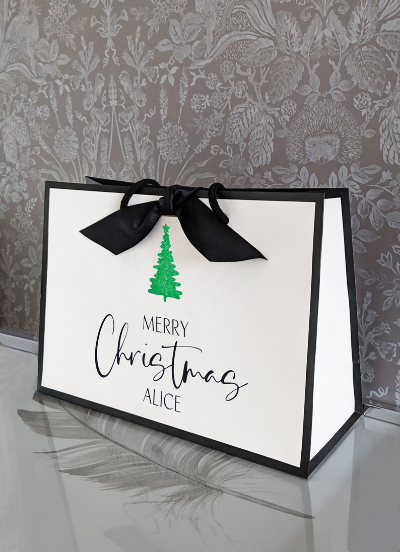 May include: White gift bag with black trim and a black ribbon bow. The bag features a green glitter Christmas tree and the words "MERRY Christmas ALICE" in black script, perfect for holiday gifting.
