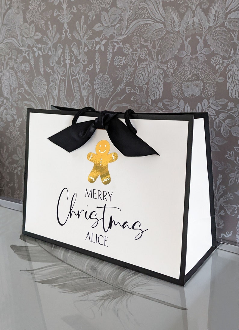 May include: White gift bag with black trim and handles, featuring a gold gingerbread man graphic. The bag has the words "MERRY Christmas ALICE" printed in black script. A black bow is tied at the top.