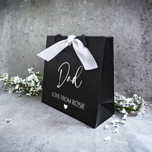 May include: Black gift bag with a white satin ribbon and the text "Dad Love From Rosie" in white lettering. A small white heart is below the text.