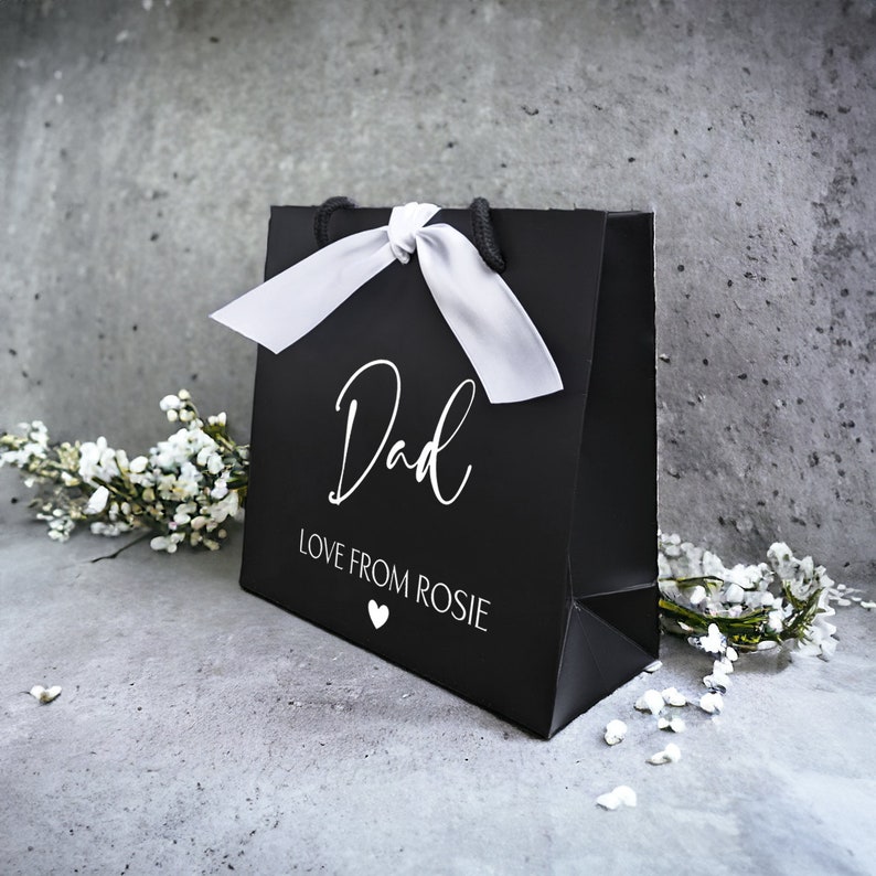 May include: Black gift bag with a white satin ribbon and the text "Dad Love From Rosie" in white lettering. A small white heart is below the text.