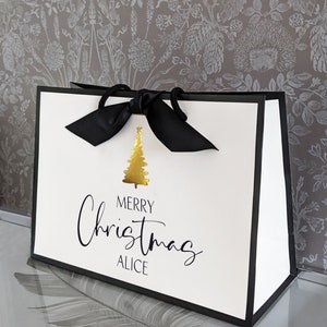 May include: White gift bag with black trim and a black ribbon bow. The bag features a gold Christmas tree graphic and the words "MERRY Christmas ALICE" in black script.