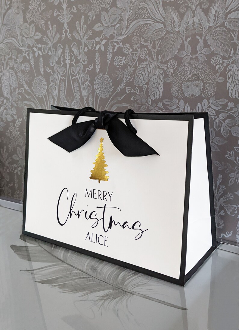 May include: White gift bag with black trim and a black ribbon bow. The bag features a gold Christmas tree graphic and the words "MERRY Christmas ALICE" in black script.
