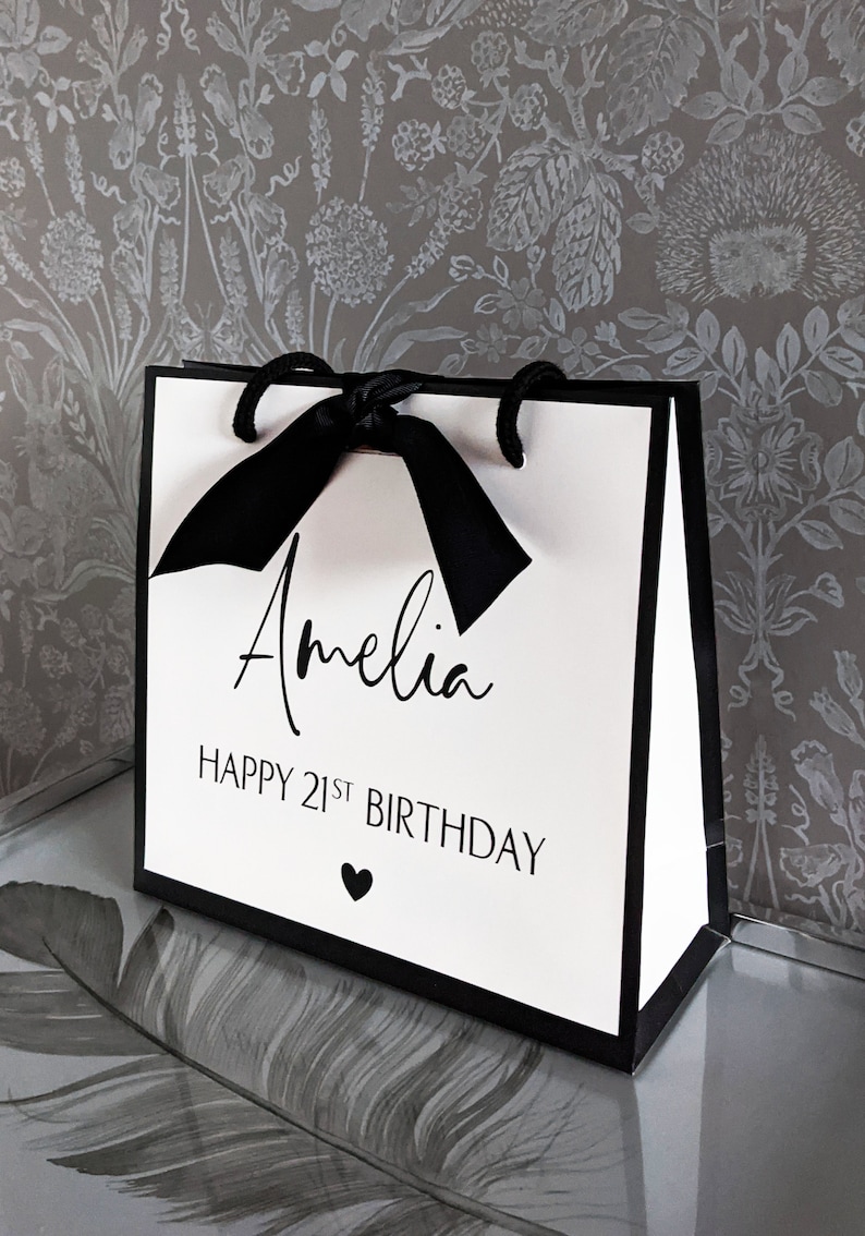 May include: A white gift bag with black trim and handles, tied with a black ribbon. The bag has the name "Amelia" and the text "HAPPY 21ST BIRTHDAY" printed on the front, along with a small heart.