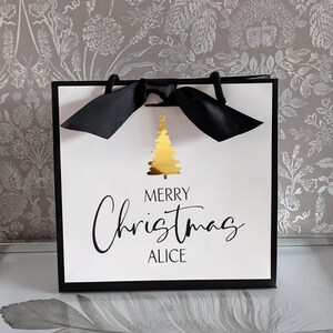 May include: A white gift bag with a black border and handles, tied with a black ribbon. The bag features a gold Christmas tree graphic and the text "MERRY Christmas ALICE" in elegant script, perfect for holiday gifting.
