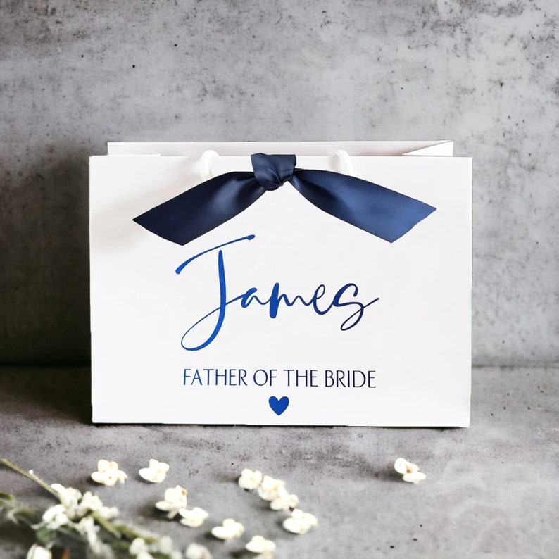 K&ouml;nnte beinhalten: Eine wei&szlig;e rechteckige Geschenkt&uuml;te mit einer dunkelblauen Satin-Schleife an der Oberseite. Auf der Vorderseite ist der Name James in blauer Schreibschrift aufgedruckt, gefolgt von dem Text FATHER OF THE BRIDE in blauen Gro&szlig;buchstaben und einem kleinen blauen Herz.