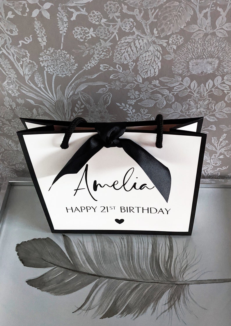 May include: White gift bag with black trim and handles, tied with a black ribbon. The bag features the name "Amelia" and the text "HAPPY 21ST BIRTHDAY" in black script. A small black heart is below the text.