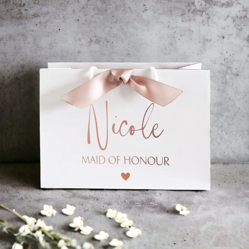 K&ouml;nnte beinhalten: Eine wei&szlig;e Geschenkt&uuml;te mit dem Namen Nicole in ros&eacute;goldener Schreibschrift, dem Text MAID OF HONOUR in ros&eacute;goldenen Gro&szlig;buchstaben, einem kleinen Herzsymbol in Ros&eacute;gold und einer ros&eacute;goldenen Satinschleife am oberen Griff.