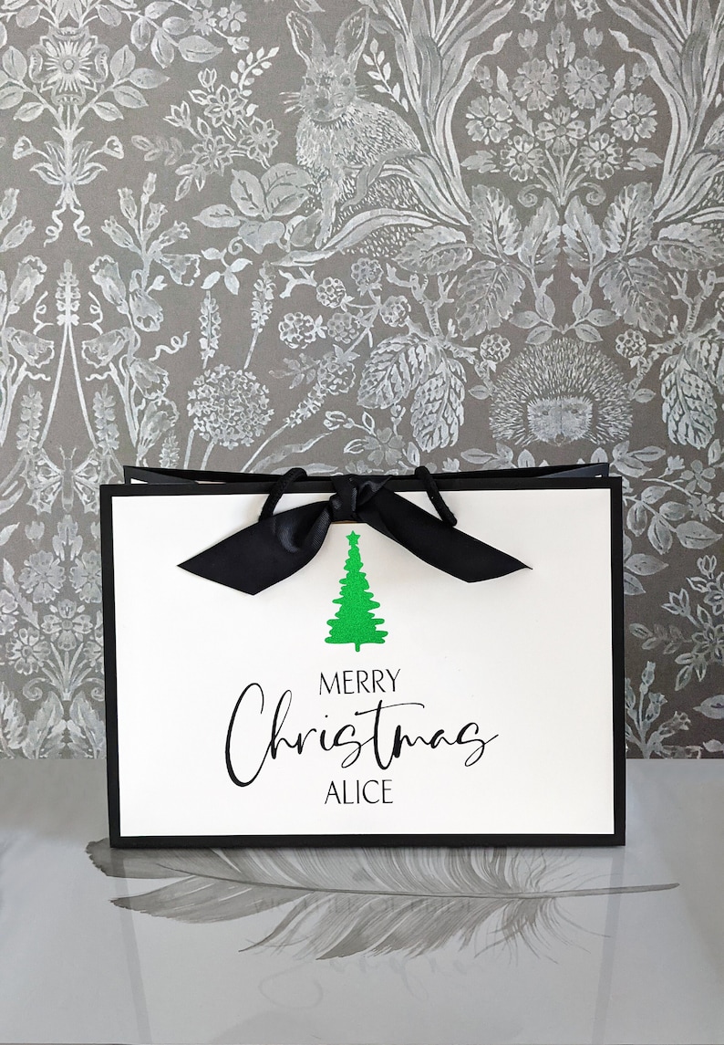 May include: A white gift bag with a black border and a black bow. The bag features a green Christmas tree graphic and the words "MERRY Christmas ALICE" in black script. The background is a gray floral pattern.