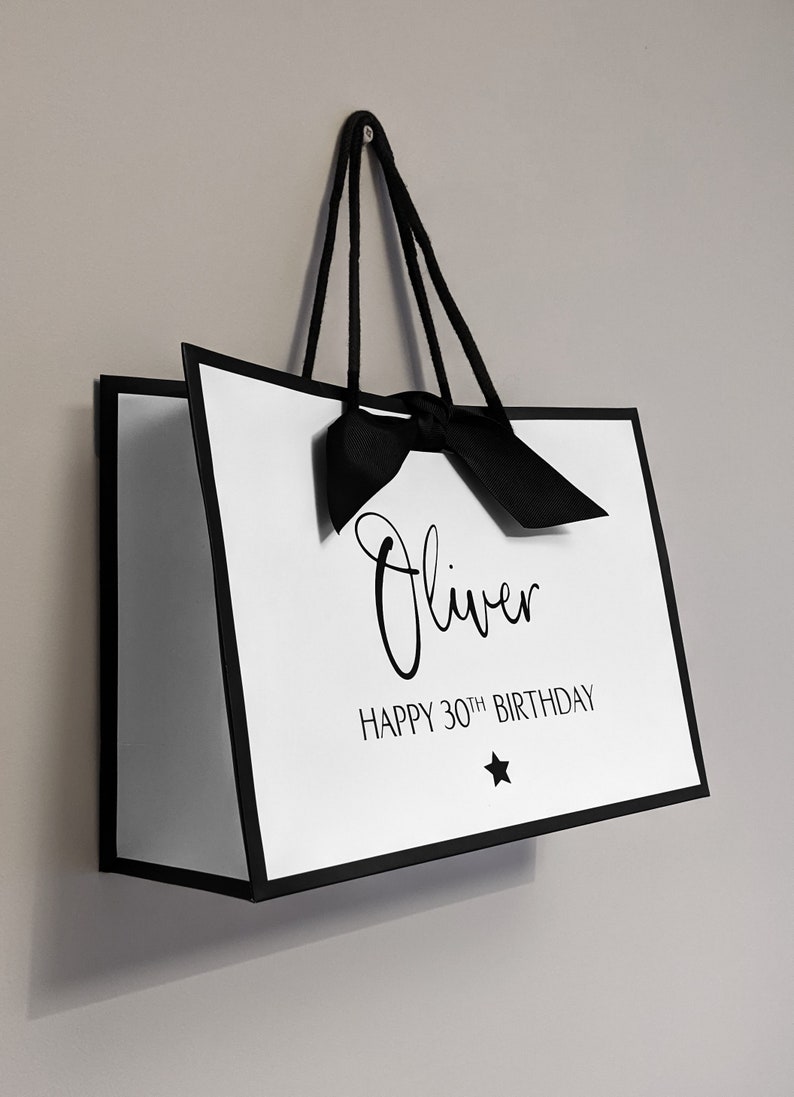 May include: White gift bag with black trim, handles, and bow. The bag has the name "Oliver" and the text "HAPPY 30TH BIRTHDAY" printed on it, along with a star graphic.