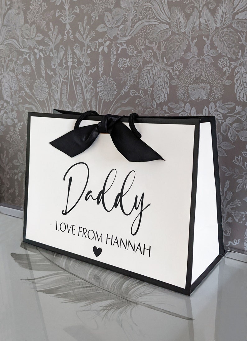 May include: A white gift bag with a black border and a black satin ribbon. The bag has the text "Daddy Love From Hannah" printed on the front.