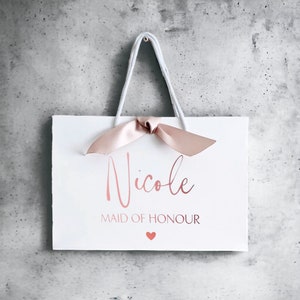 K&ouml;nnte beinhalten: Eine wei&szlig;e Geschenkt&uuml;te mit wei&szlig;en Kordelgriffen und einer altrosa Satinschleife. Die T&uuml;te ist mit dem Namen Nicole in ros&eacute;goldener Schreibschrift bedruckt, gefolgt von dem Text MAID OF HONOUR in ros&eacute;goldenen Gro&szlig;buchstaben und einem kleinen rosa Herz.
