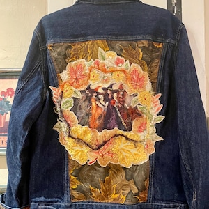 May include: A dark blue denim jacket featuring a large, circular fabric appliqué on the back. The appliqué depicts three figures in witch hats surrounded by a wreath of embroidered autumn leaves in shades of orange, yellow, and red.