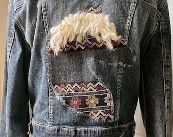 Könnte beinhalten: Jeansjacke in Blau mit einem einzigartigen Stickdesign auf dem Rücken. Das Design zeigt ein weißes, strukturiertes Element und farbenfrohe Blumen- und geometrische Muster. Die Jacke ist mittelblau.