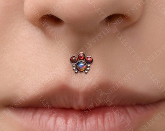 May include: A silver-toned philtrum piercing jewelry piece shaped like a small paw print. The design features four circular, iridescent red-toned stones set in a metallic frame, positioned directly above the center of the upper lip.