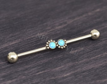 Puede incluir: Una pieza de joyería corporal tipo barra industrial metálica con dos piedras redondas de color azul claro similares al ópalo en el centro. Las piedras están rodeadas de pequeñas cuentas metálicas y la barra termina en esferas pulidas.