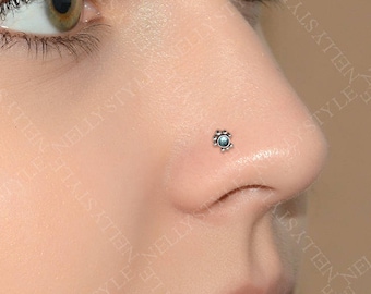 May include: A small, circular nose stud featuring a central blue stone surrounded by a scalloped metal border. The jewellery is positioned on the side of the nostril, showcasing a detailed, flower-like design with a polished silver-toned finish.