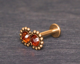 May include: Gold body jewelry with two orange gemstones set in a beaded design.