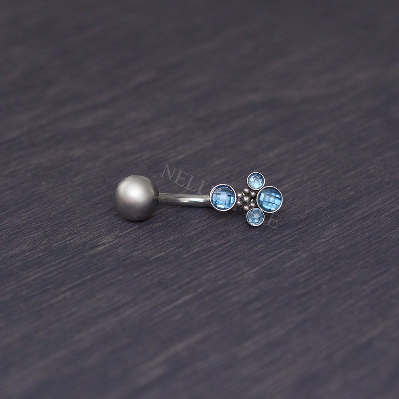 May include: A silver belly button ring with a cluster of four light blue gemstones. The ring has a small, round ball on one end and a decorative arrangement of gems on the other. The gems are set in a silver metal.
