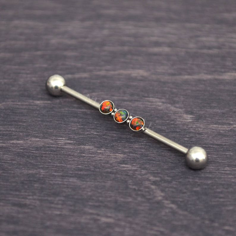 Puede incluir: Una barra de plata con cuatro piedras preciosas que parecen &oacute;palos en tonos naranja y verde. La barra es una joya de piercing corporal.