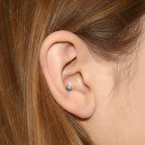 May include: A close-up of a person's ear with a gold ear piercing featuring a blue gemstone and small gold beads.