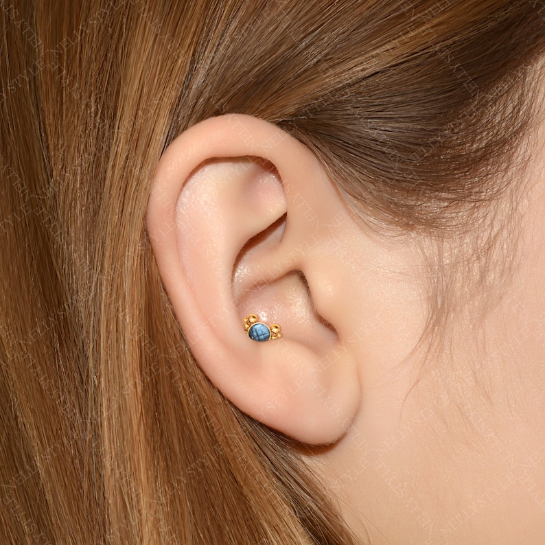 May include: A close-up of a person's ear with a gold ear piercing featuring a blue gemstone and small gold beads.