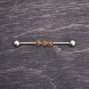 Puede incluir: Una barra de barbell plateada con tres piedras que parecen &oacute;palos en fila. Las piedras son una mezcla de naranja, rojo y negro.