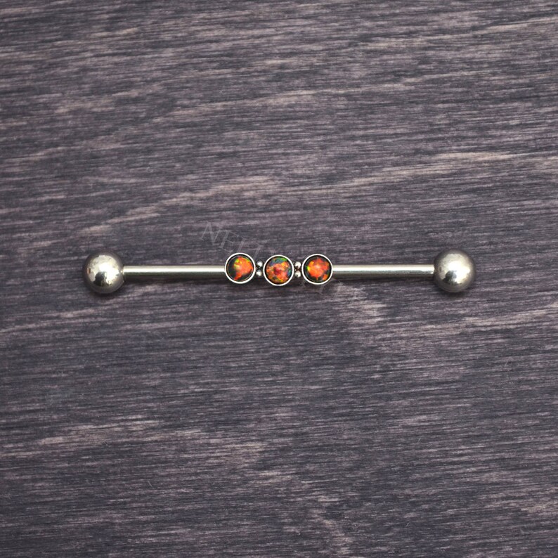 Puede incluir: Una barra de barbell plateada con tres piedras que parecen &oacute;palos en fila. Las piedras son una mezcla de naranja, rojo y negro.