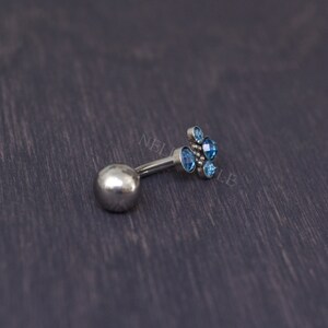 May include: A silver-colored belly button ring with a cluster of light blue gemstones. The ring features a small ball on one end and a decorative floral design on the other. The gemstones are round and faceted.