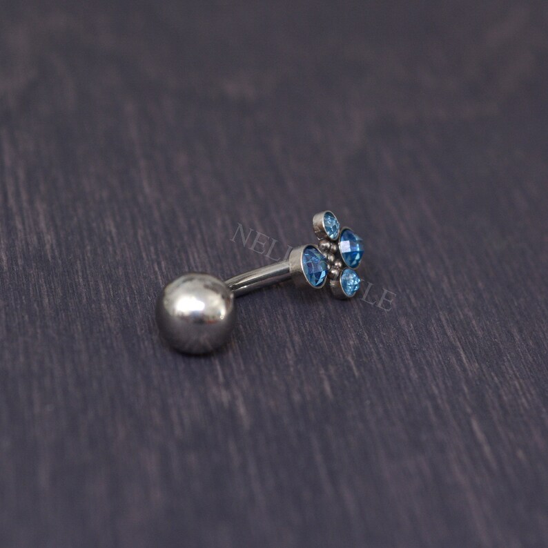 May include: A silver-colored belly button ring with a cluster of light blue gemstones. The ring features a small ball on one end and a decorative floral design on the other. The gemstones are round and faceted.