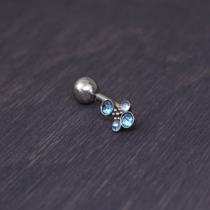 May include: A silver-colored helix earring with a cluster of five light blue gemstones. The earring has a small, round ball on one end and a decorative arrangement of gemstones on the other. The gemstones are round and vary slightly in size.