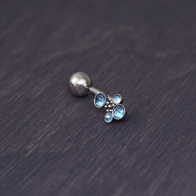 May include: A silver-colored helix earring with a cluster of five light blue gemstones. The earring has a small, round ball on one end and a decorative arrangement of gemstones on the other. The gemstones are round and vary slightly in size.